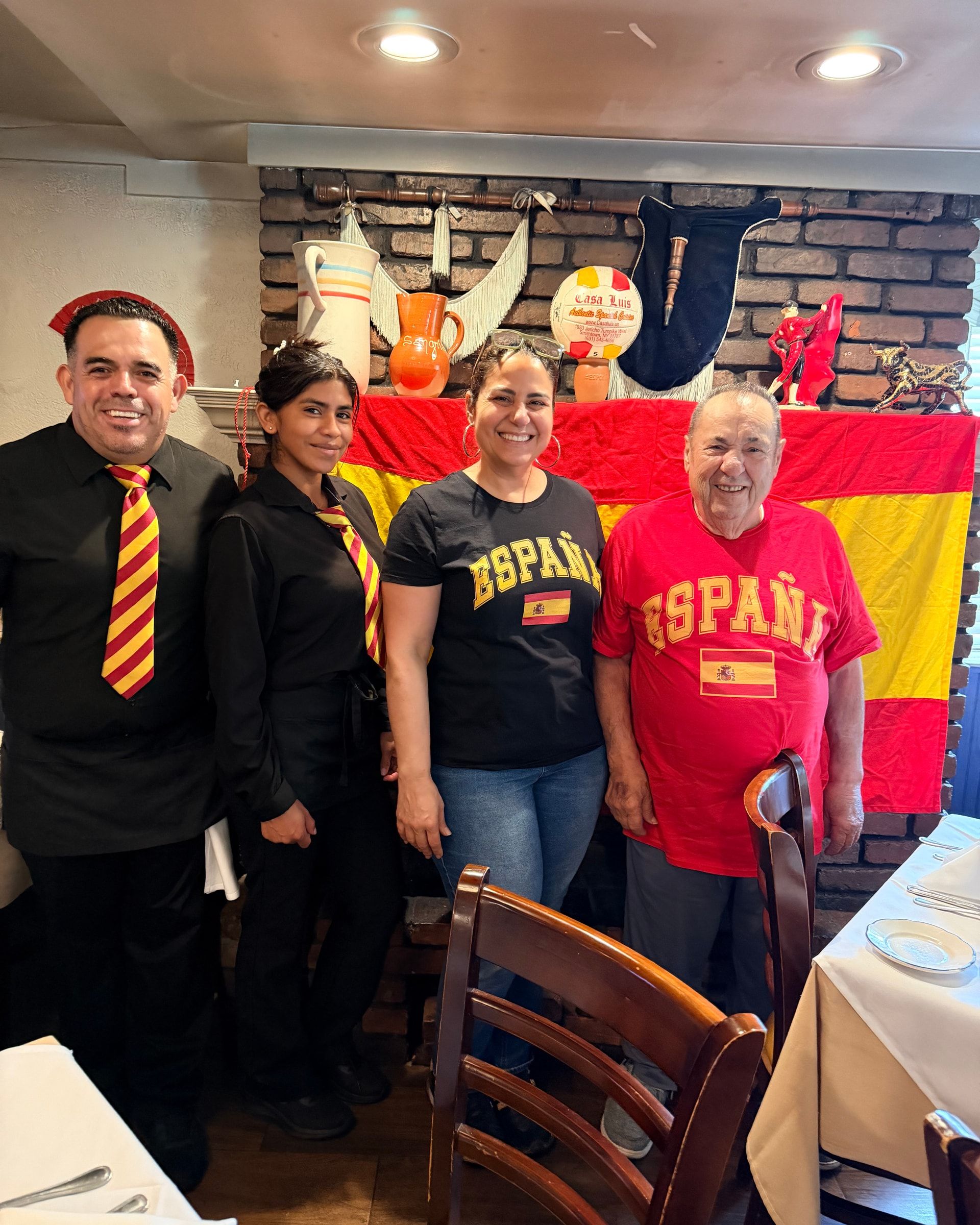 Delia and Luis in España shirts with Casa Luis staff by the fireplace and the Spanish flag